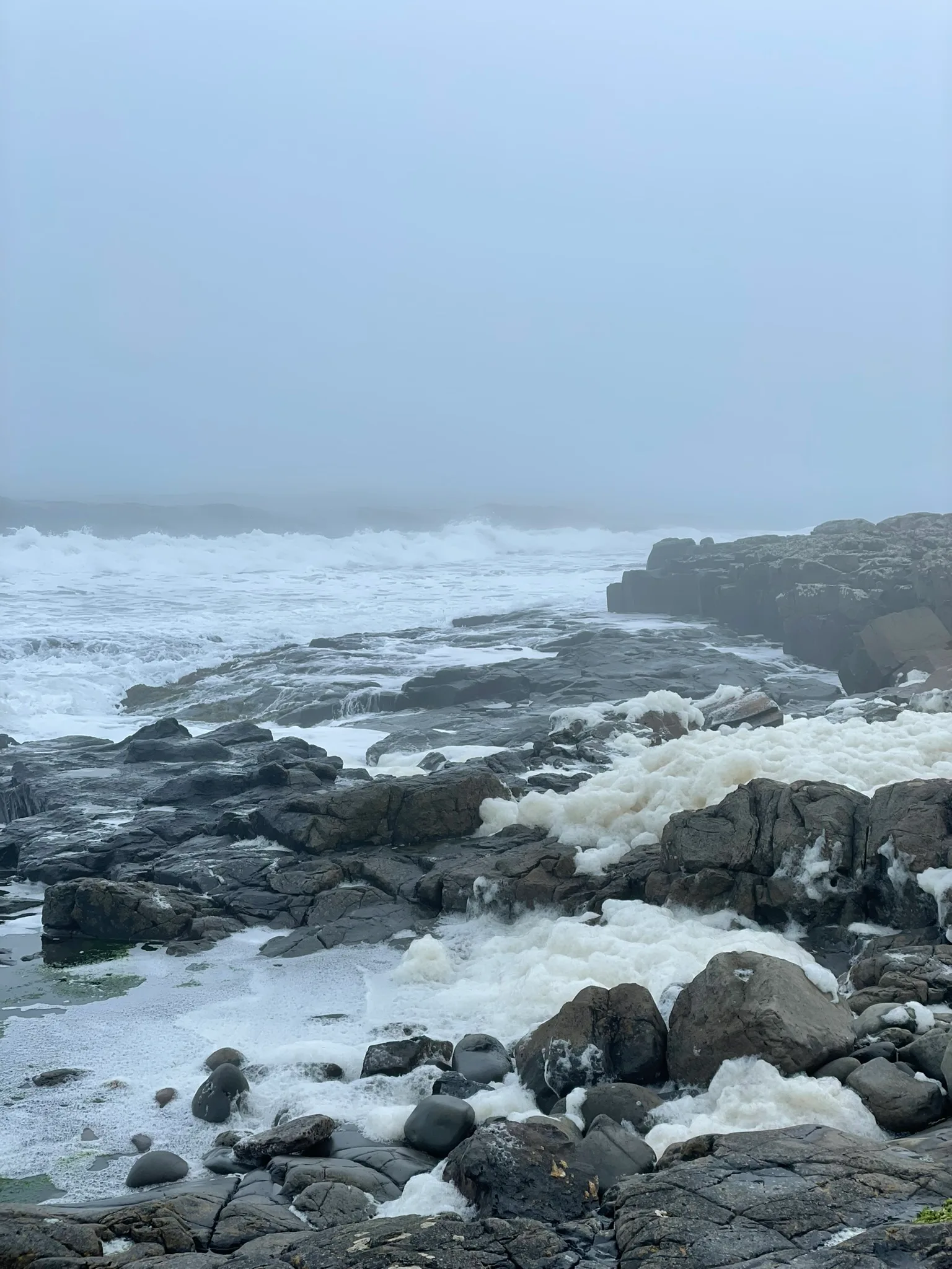 Dramatic waves crashing against weathered rocky shore shrouded in mystical fog, embodying the power and mystery of coastal spirituality